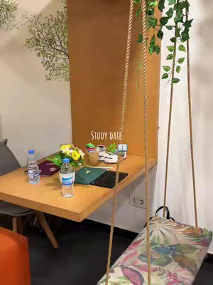 The image shows a cozy study setup at a cafe. A wooden table holds a laptop, some water bottles, a notebook, and a bouquet of flowers. A swing with a patterned cushion is in the background. The words 'STUDY DATE' are written on the wall. The color palette includes natural wood tones, white, and splashes of green from the plants.