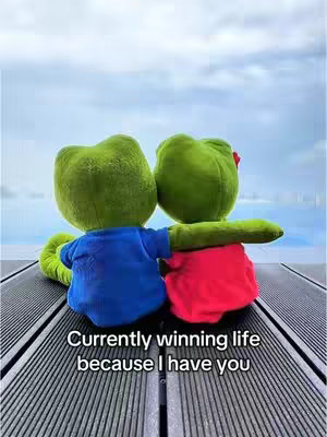 The image shows two plush frog toys from the back, snuggled together on a wooden surface. The frog on the right has a red bow on its head, and its arm is wrapped around the other frog. Behind them is a cloudy sky. Text overlayed at the bottom of the image reads 'Currently winning life because I have you'.