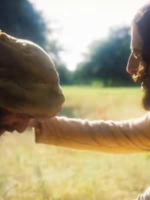 An outdoor scene featuring a man in brown robes reaching a hand towards another person's head, presumably for a blessing. The background suggests a field setting. The lighting is soft, creating a serene and spiritual atmosphere.