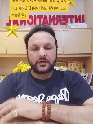 A man with dark hair and a beard is in a room. He is wearing a dark blue t-shirt with white writing. There is text in a yellow box above his head. Behind him is a shelf with various items, including two framed pictures and some books. The background is a mix of brown and light colors.