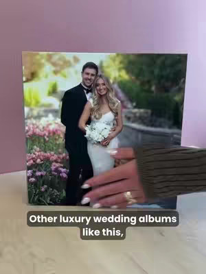A photo printed on a canvas. It features a wedding photo of a man in a black suit and a woman in a white wedding dress, holding a bouquet. They are standing in front of a floral setting. A hand and arm, wearing a green sweater, is holding the canvas. The background is a mix of pink and white.