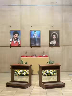 The image displays a beige wall with three framed portraits hanging on it. The bottom center is a light wooden structure with reflection and a floral arrangement. In front of it, placed on a lower wooden structure.  The portraits depict three individuals, presumably saints, as the caption suggests. There is a decorative element above the center picture. Two lectern structures with floral arrangements sit in front of the center.