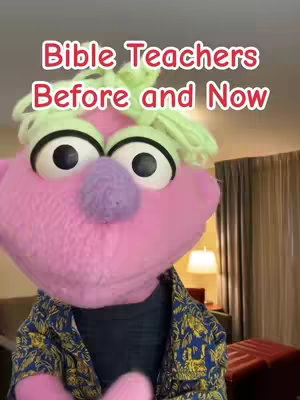 A pink puppet with large eyes and a green, curly hairstyle is centered in the frame. The puppet is wearing a patterned shirt and a black undershirt. The background has a blurry hotel interior.