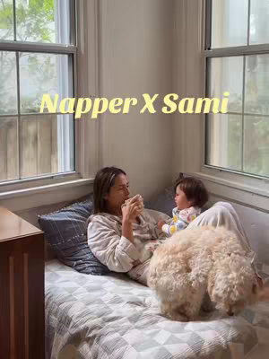 A woman and a baby are lying on a bed with a dog in the image. The woman is wearing a robe and drinking from a cup, looking towards the baby. The baby is sitting up in front of her. A fluffy white dog is in the front portion of the bed and both look out of the image. The scene is lit with natural light from two windows.