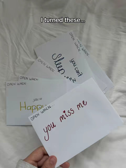A hand holding multiple white envelopes, each with handwritten messages like "Sleep" and "you miss me". The envelopes are arranged on a wrinkled white bedsheet.
