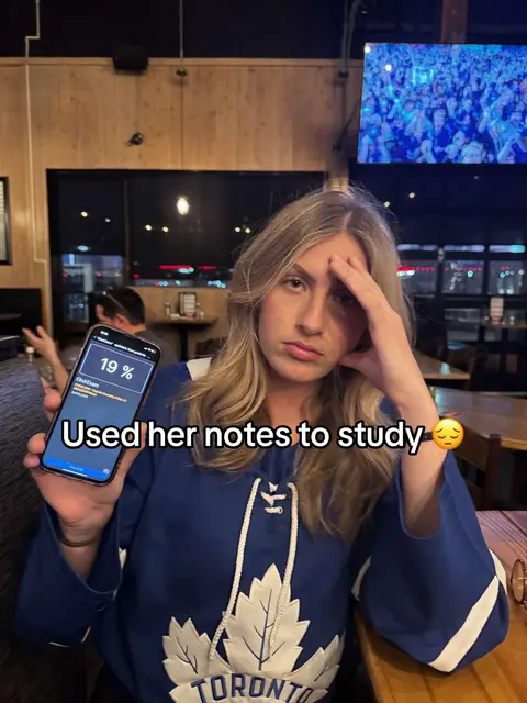 A woman in a blue hockey jersey sits at a table, holding a phone displaying a low percentage reading. She has her hand on her forehead and a somewhat pained expression on her face, suggesting frustration or disappointment. The background includes a blurry restaurant interior with other patrons.