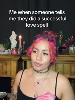 A medium shot featuring a woman with pink hair, wearing a black top, and several necklaces. She is in front of a decorative wooden cabinet that has a candle and statues on top of it. Text overlays the top of the image.
