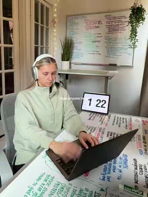 A young woman is seated at a desk in front of a whiteboard covered in notes. She is wearing headphones and looking down at a laptop. Her hands are blurred, suggesting she is actively typing. Behind her is a bright window and a plant. A digital clock displaying '1 22' is visible. There are notes and writings on her desk. The color scheme is bright and the lighting is natural.
