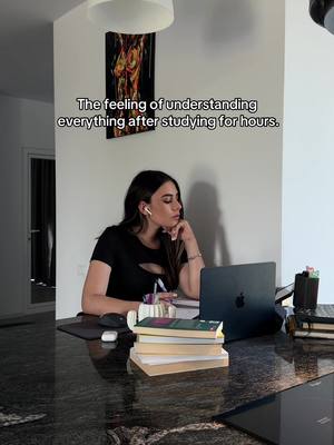 A young woman is seated at a kitchen counter, absorbed in study. She is wearing a black shirt and has headphones in, with a laptop open in front of her and stacks of books nearby. The background includes a modern kitchen with a minimalist painting on the wall.