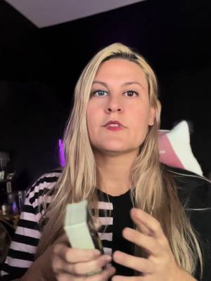 A woman with long blonde hair, wearing a black and white striped shirt and a black top, is centered in the frame. Her expression is neutral as she looks towards the camera. Her hands are up, seemingly gesturing and a light teal object she is grasping. The background is a gradient of dark grey and black. A pink and white chair back is seen on her right.