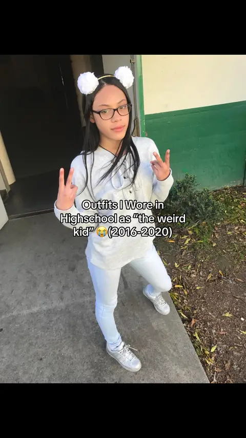 A person stands in front of a building, wearing a white outfit and a pair of glasses. They are making a peace sign with their fingers. Text overlayed says "Outfits I Wore in Highschool as 'the weird kid' (2016-2020)"