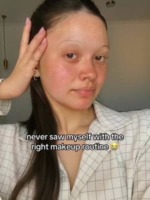 A young woman with fair skin and dark hair is shown in a close-up, likely a selfie. She is in a well-lit room, wearing a white and gray checkered button-down. The main focus is on her face, with her hand gently touching her face near her temple. Below her, a text overlay reads "never saw myself with the right makeup routine".