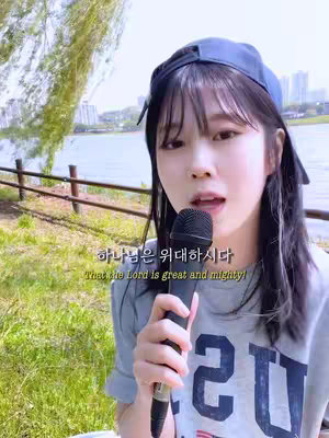 A girl is holding a black microphone and singing, the background has green nature and a lake. There is a text overlay in the image.