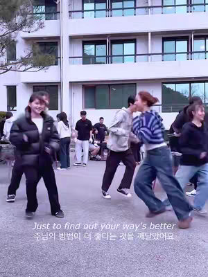 A group of young people are dancing outdoors in front of a building with white walls and multiple windows. The foreground features people dancing with energetic movements while wearing casual clothes. Text is overlaid on the lower portion of the image in both English and Korean text, and the overall scene is brightly lit, suggesting an upbeat and positive environment.