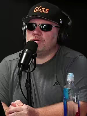 A man wearing a black cap, sunglasses, and headphones speaks into a microphone. A bottle of water is visible in front him.