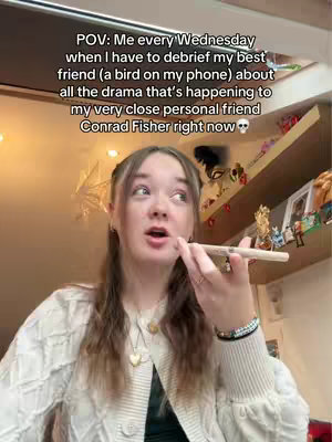 A young woman is shown in a selfie style shot. She is talking with clear emotion with a pen in her hand and an open mouth. The background shows some items displayed on some shelves.