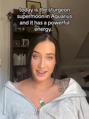 A woman with long dark hair poses in what looks like a home interior, with text overlayed at the top. She is wearing a grey zip-up, and has a silver necklace with a green pendant. In the background is a dark bookshelf and a mirror.