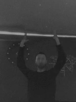 A black and white photo shows a man with arms raised overhead. The background appears to be a dark wall. The positioning of his arms and hands creates a symmetrical, almost posed arrangement. His face is somewhat blurry, but he appears to be looking towards the viewer. Above him, and extending across the top, is a horizontal light fixture or a band of light. Behind him, two stylized circular arrangements of points are visible on the wall.