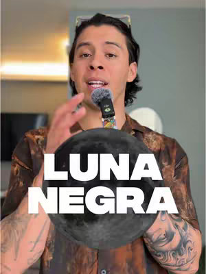A man with dark hair and sunglasses perched on his head stands in front of a background with light and shadow. He is holding a microphone and gestures with his hands. In the foreground is a large black sphere with the white text "LUNA NEGRA". He wears a brown shirt, tattoos, and his expression is animated.