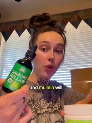 A woman with dark hair in a bun, holds a dropper bottle near her face. The label is bright green and shows the words 'Organic' and 'Mullein'. A partial view of a white container is visible. Background curtains are open.