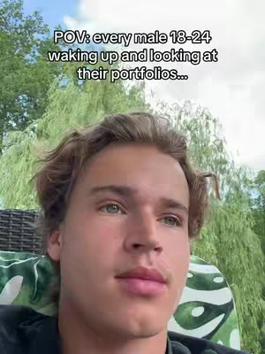 A young man with light skin and wavy brown hair is the primary focus. He has green eyes, and is looking at the camera with a neutral expression. The background includes lush green foliage and a glimpse of the sky, suggesting an outdoor setting.
