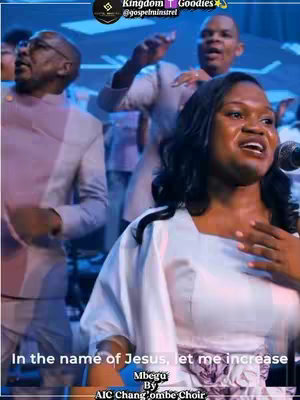 A group of people are singing in what appears to be a church setting. A woman in a white dress sings into a microphone as others around her sing and gesture as well.