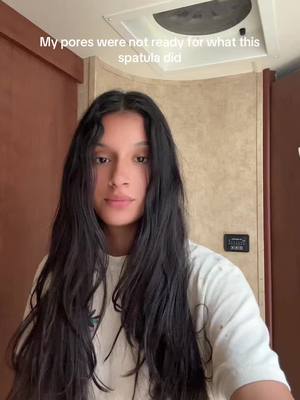 A woman with long, wavy black hair stares at the camera in a selfie-style shot. The background shows a light-colored wall and a portion of a ceiling fixture. She's wearing a white t-shirt, and the overall lighting appears natural. Text overlayed on the image reads, 'My pores were not ready for what this spatula did.'