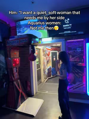 A woman stands in what appears to be an arcade or game room, near a boxing game. Text overlays on the image, with the creator's back to the camera.