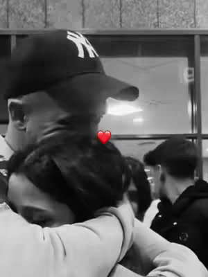 A black and white image features two people embracing. The person in the foreground has their head resting on the shoulder of the other, who wears a baseball cap. A small red heart is superimposed between them.