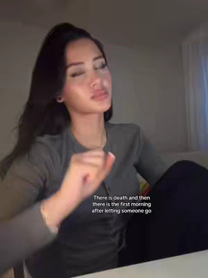 A woman with dark hair, wearing a gray long-sleeve shirt, is seated in front of a neutral background. She has her eyes closed and face tilted upwards slightly; one hand is raised, fingers partially curled, and the other arm is bent at the elbow, resting on their leg. Text, in a white sans-serif font, overlays the image at the bottom: 'There is death and then there is the first morning after letting someone go'. The overall lighting is soft, suggesting an indoor setting.