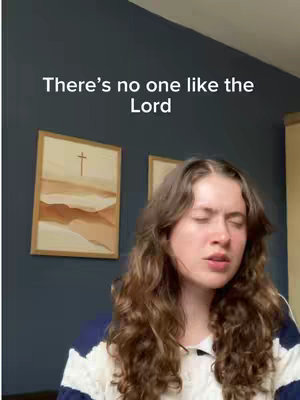 A young woman with long, curly brown hair sings with her eyes closed, illuminated by soft lighting. A sign with a cross and a second, obscured sign are visible on the background while she is wearing a white and blue striped top. The text overlay reads 'There's no one like the Lord'.