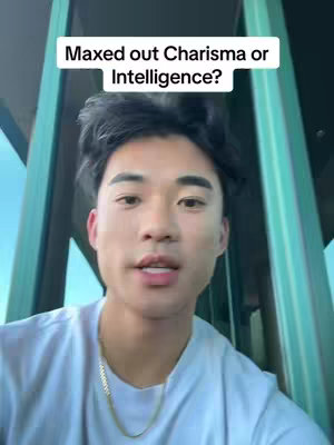A close-up shot of a young man with dark hair and a light complexion. He is looking towards the camera with an open mouth. Above him, a text overlay asks, "Maxed out Charisma or Intelligence?" The background is a bit blurry, with green and blue tones. He's wearing a white t-shirt and a gold necklace.
