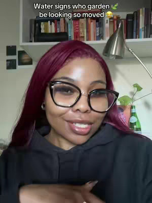 A medium shot of a Black woman with dark burgundy hair, large black-framed glasses, and a black hoodie. She is looking towards the camera and smiling. The background is softly blurred, including a shelf with books and some decorative items. The lighting is soft and natural.