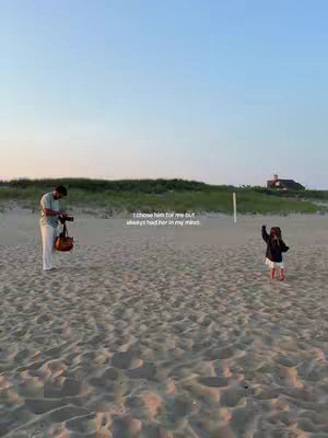 A wide shot captures a man and a young child on a beach at dusk. The man, standing on the left, is holding a camera and a satchel. The child, in the right of the frame, is facing the man with their arms outstretched. The scene is bathed in soft, warm light, suggesting either sunrise or sunset. The beach itself appears to be sandy and expansive, while a grassy dune and a house are in the background.