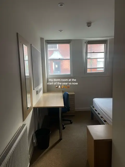 An image of a dorm room, viewed from the entrance. The room is sparsely furnished. There's a desk with a blue office chair, a bed with a white cover, a small bedside table, and a black trash can. The walls are white with two windows on the right side of the frame. The room appears to be lit by natural light.