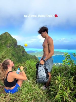 A scenic outdoor shot featuring a couple in a mountainous landscape. A woman in a black top and blue shorts sits on the left, looking up. A man, shirtless and wearing grey shorts, stands on the right, facing her. The background showcases a clear blue sky, green mountains, and turquoise water.