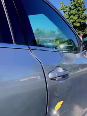 The image shows a close-up of a silver car, focusing on the front passenger side. The window is tinted, reflecting a blurred view of trees and sky. A decorative yellow feather sticker is placed near the car door's bottom, reflecting light.