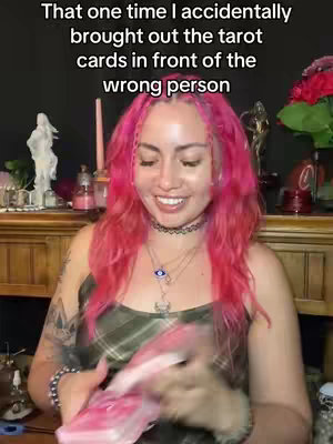 A woman with pink hair smiles while holding a deck of tarot cards. The background is slightly blurred but suggests a room with various items on shelves and surfaces. Overlay text at the top reads, "That one time I accidentally brought out the tarot cards in front of the wrong person."