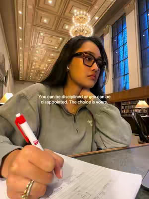 The cover image features a young woman in a library, presumably from Columbia University, as suggested by the hashtags. She's wearing glasses and a casual gray sweatshirt and is writing on a piece of paper with a pen. The background is blurred but features columns, possibly a chandelier, and a large window bringing in soft, natural light. Overlayed is text that reads 'You can be disciplined or you can be disappointed. The choice is yours.'