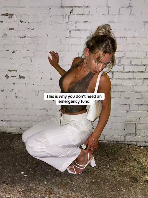 A woman in a brown top and white pants is kneeling in front of a white brick wall, with a white purse, high heels, and a text overlay. A sign says "This is Why you don't need an emergency fund."