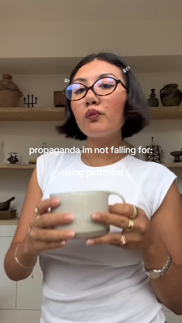 A woman with short, dark hair and glasses is holding a mug. She's wearing a white top and several rings. The background shows what appears to be a kitchen. Text overlays the image.