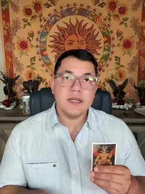 A man wearing glasses and a striped shirt sits in front of a decorative tapestry and holds a tarot card aloft. Behind him are decorative items on a shelf.