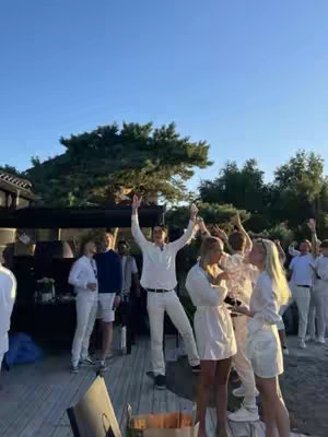 A group of people in a sunny outdoor setting, likely a party or social gathering. People are standing on a wooden deck, with a man in the center raising his arms in celebration. Many are dressed in white attire. The background includes trees and a clear blue sky, suggesting a daytime event.