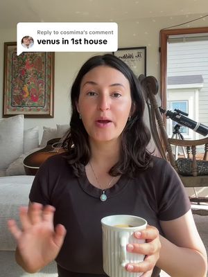 A woman is centered in the frame, looking directly at the camera and speaking. She is holding a white mug in her right hand. Behind her is a warm-toned room with a window to her right and art on the wall behind her, along with an acoustic guitar and a sofa to the left.