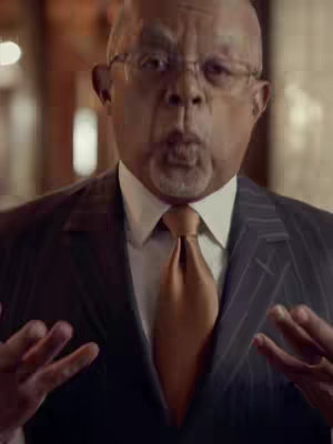 A medium shot of a man in a suit with hands up and an expression, as if he is about to speak or making a point.