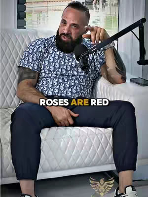 A man with a dark beard and short hair sits on a white quilted sofa with a microphone in front of him. The image features an overlay of text that reads "ROSES ARE RED".