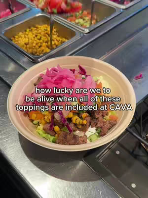 A close-up shot of a bowl filled with various food items, including what appears to be a grain base, colorful toppings like corn and pickled onions, and meat. The bowl is beige, set against a metallic background and food prep stations.