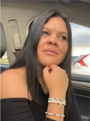 A young woman with long dark hair is seated in the passenger seat of a car, gazing off to the side with a pensive expression. She's wearing a black off-the-shoulder top and multiple bracelets on her wrist. Her hand is raised to her chin. Background elements include the interior of the car and a glimpse of the outside environment.