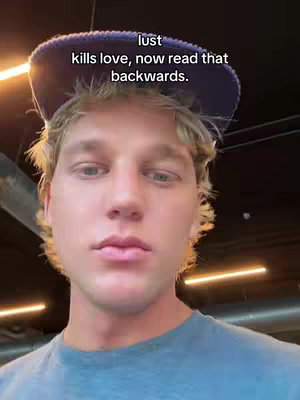 The cover image features a close-up of a young man with fair skin and blond hair set against a blurred background. He wears a blue hat and a blue shirt. Overlayed text in the upper part of the image reads, "lust kills love, now read that backwards." The lighting is warm, and the composition focuses intensely on his face.