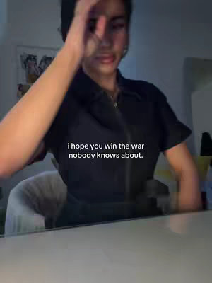 A person is shown from the chest up, with their face slightly out of focus and towards the camera. They are wearing a dark colored button up with a zipper down the middle. Their arm is up in front of their face, touching their head. The background is dark, and there is some text in white font at the bottom of the person.